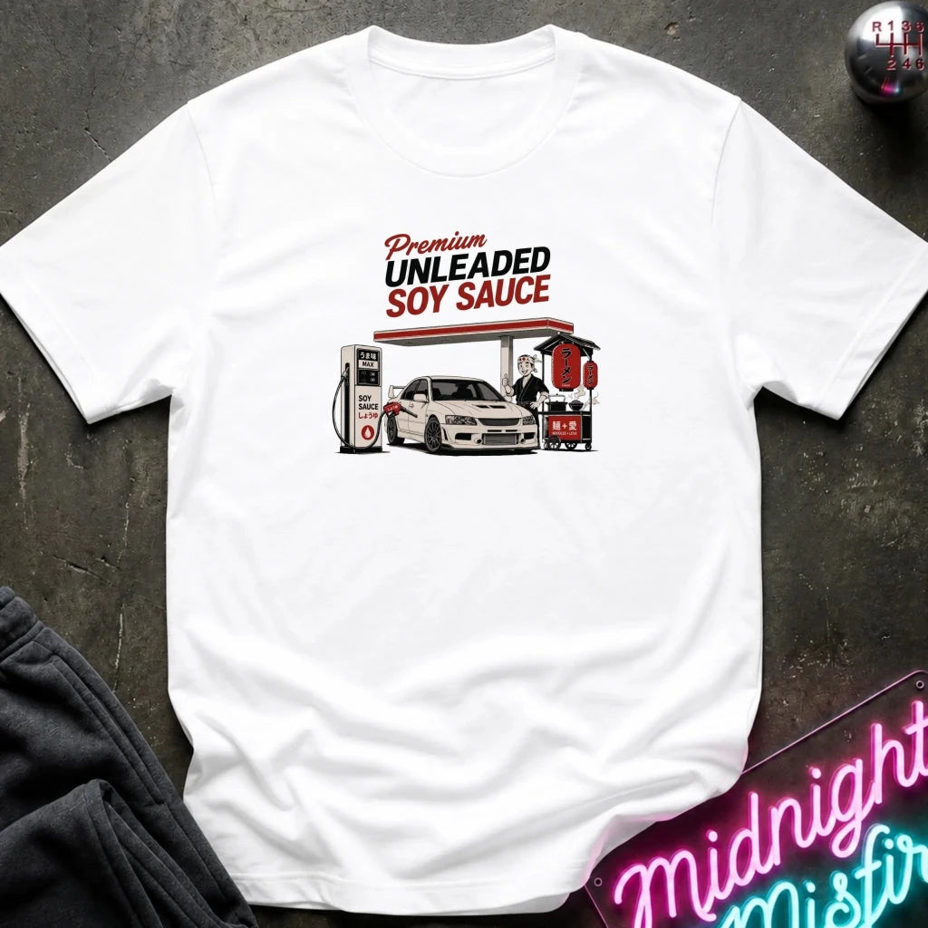 A white short-sleeve graphic T-shirt laid flat on a dark textured surface. The shirt features a retro-style gas station illustration with the text 'Premium UNLEADED SOY SAUCE' above a white car and fuel pumps.