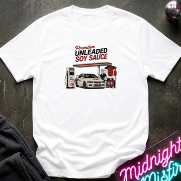 A white short-sleeve graphic T-shirt laid flat on a dark textured surface. The shirt features a retro-style gas station illustration with the text 'Premium UNLEADED SOY SAUCE' above a white car and fuel pumps.