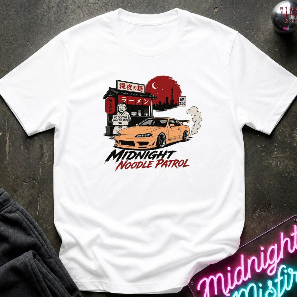 A white short-sleeve graphic T-shirt laid flat on a dark textured surface. The shirt features a printed illustration of an orange sports car in front of a ramen shop with a red sun and smoke, with the text 'MIDNIGHT NOODLE PATROL' below.