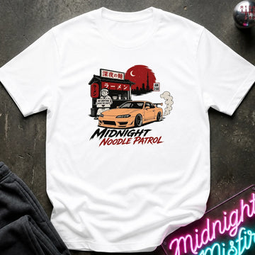 A white short-sleeve graphic T-shirt laid flat on a dark textured surface. The shirt features a printed illustration of an orange sports car in front of a ramen shop with a red sun and smoke, with the text 'MIDNIGHT NOODLE PATROL' below.