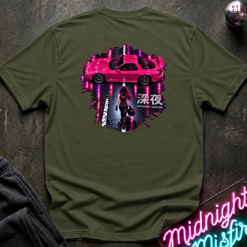 An olive green t-shirt laid flat on a dark textured surface, featuring a large graphic print of a pink sports car on a road with a woman walking underneath and Japanese text. A neon sign and folded dark clothing are partially visible in the corners.