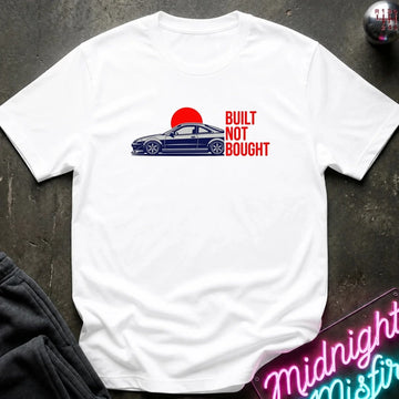 A white short-sleeve t-shirt laid flat on a dark textured surface, featuring a graphic of a blue sports car in front of a red sun with the text 'BUILT NOT BOUGHT' in red. A neon sign and dark pants are partially visible as props.