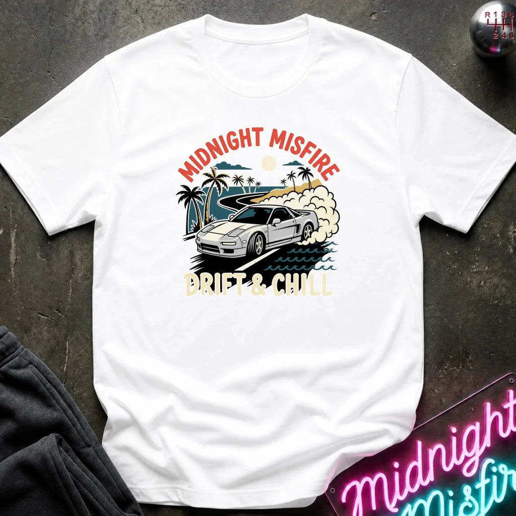 A white short-sleeve graphic T-shirt laid flat on a dark textured surface. The shirt features a retro-style print of a white sports car drifting on a beach road with palm trees, waves, and the text 'MIDNIGHT MISFIRE' and 'DRIFT & CHILL' in red, beige, and blue tones.