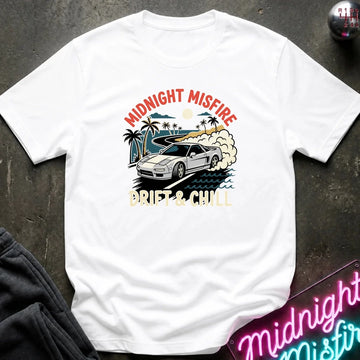A white short-sleeve graphic T-shirt laid flat on a dark textured surface. The shirt features a retro-style print of a white sports car drifting on a beach road with palm trees, waves, and the text 'MIDNIGHT MISFIRE' and 'DRIFT & CHILL' in red, beige, and blue tones.