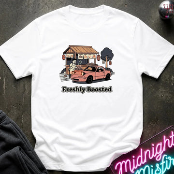 A white short-sleeve graphic t-shirt laid flat on a dark textured surface. The graphic shows a pink sports car parked in front of a small shop with the text 'Freshly Boosted' underneath.