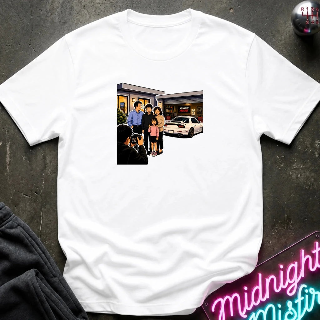 A white short-sleeve crewneck t-shirt laid flat on a dark textured surface, featuring a rectangular graphic print of a family standing outside a building taking a photo.
