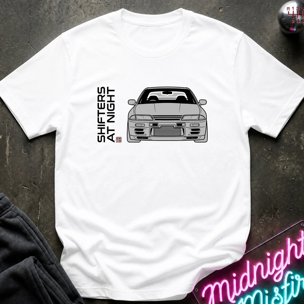 A white short-sleeve t-shirt laid flat on a dark textured surface, featuring a black line art graphic of a sports car and the vertical text 'SHIFTERS AT NIGHT' with a small red and blue flag detail. A neon sign and dark clothing are partially visible as props.
