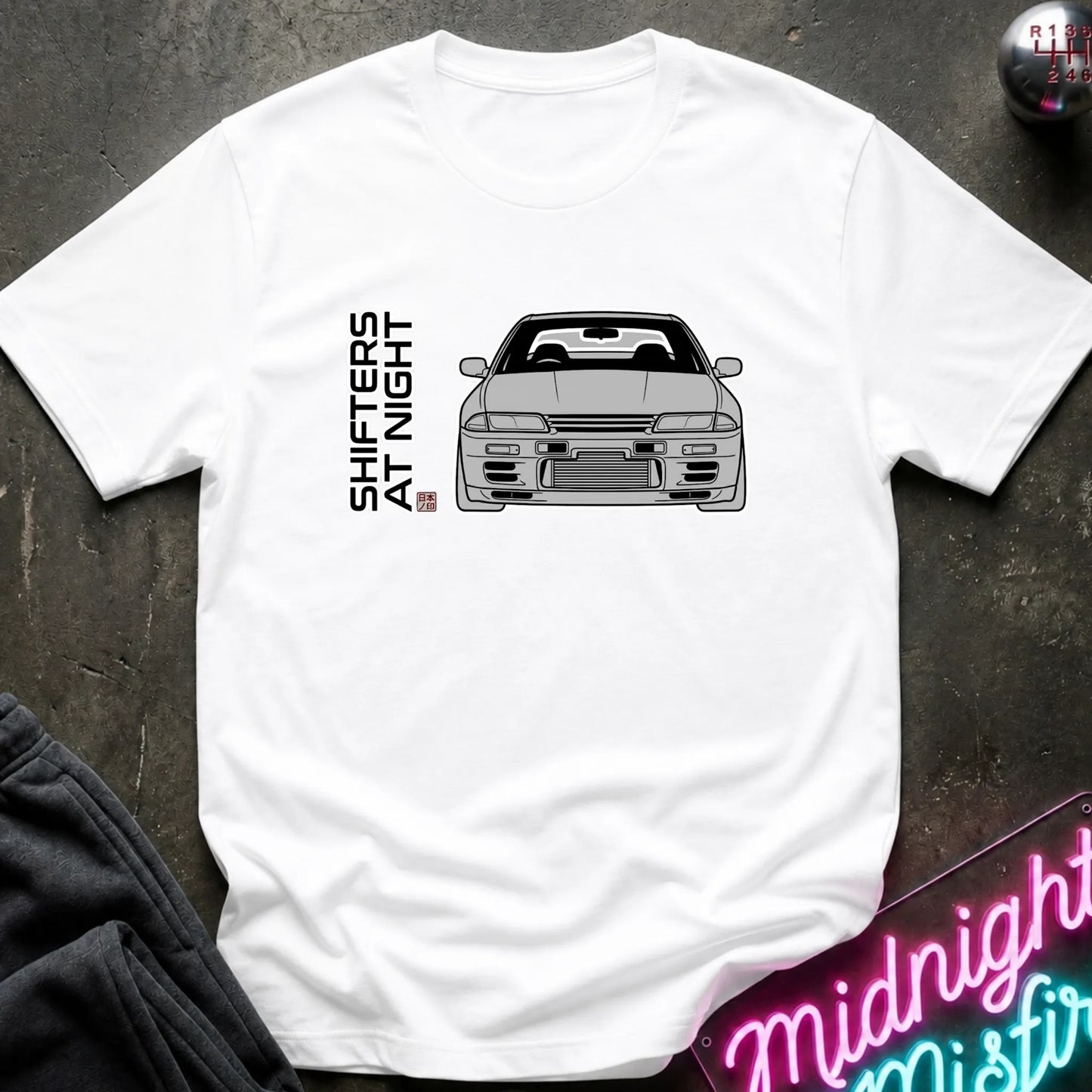 A white short-sleeve t-shirt laid flat on a dark textured surface, featuring a black line art graphic of a sports car and the vertical text 'SHIFTERS AT NIGHT' with a small red and blue flag detail. A neon sign and dark clothing are partially visible as props.
