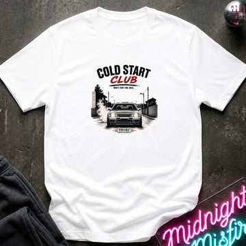 A white short-sleeve crewneck t-shirt with a graphic print of a white station wagon with a roof rack, seagulls, and a small building, laid flat on a dark textured surface.