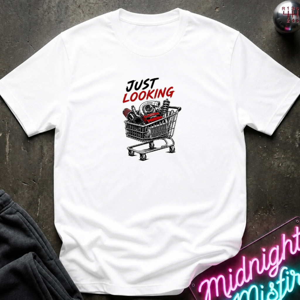 A white short-sleeve t-shirt laid flat on a dark textured surface, featuring a graphic print of a shopping cart filled with items and the text 'JUST LOOKING' in black and red. Partially visible props include dark pants in the lower left and a neon-style sign in the lower right.