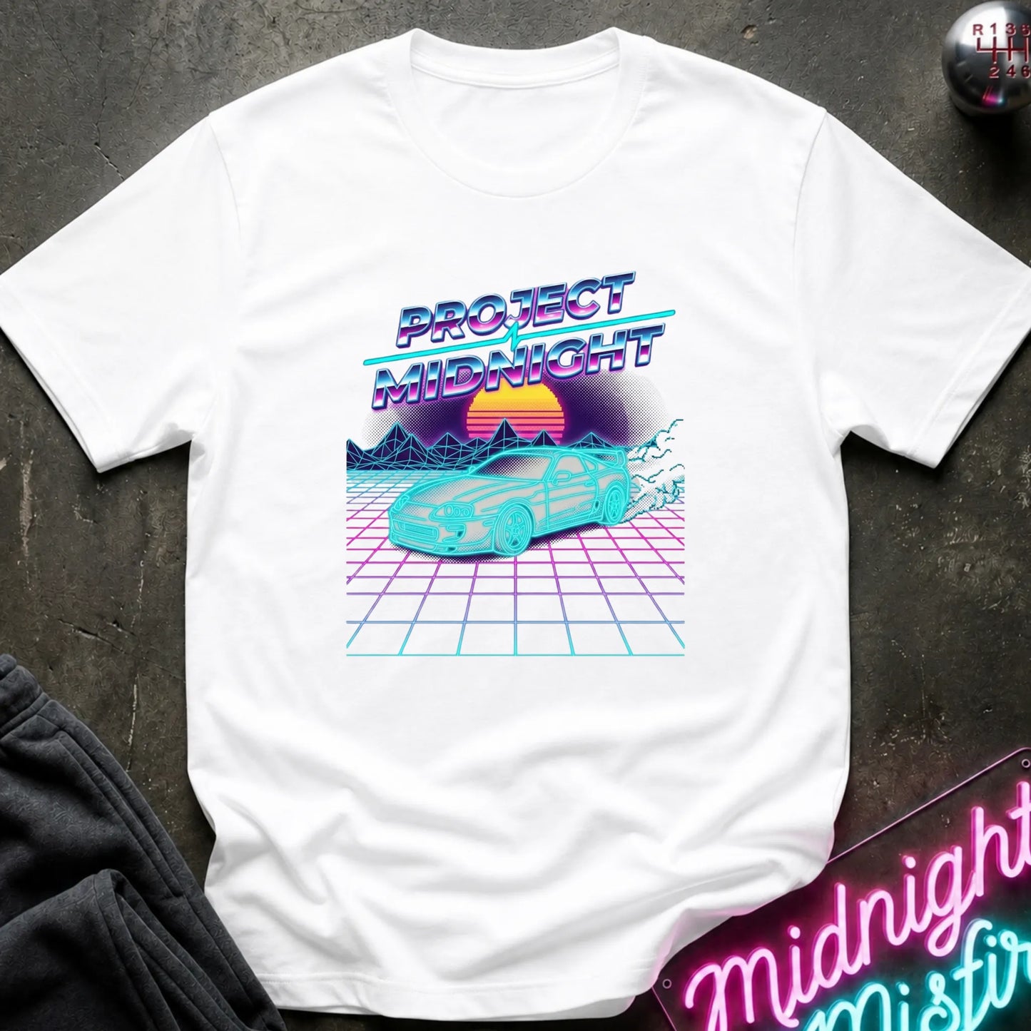 A white short-sleeve t-shirt laid flat on a dark textured surface, featuring a colorful retro 1980s-style graphic with the text 'PROJECT MIDNIGHT,' a cyan car, and a pink-to-yellow sunset over a grid pattern. Partial dark clothing and a neon sign are visible at the edges as styling props.