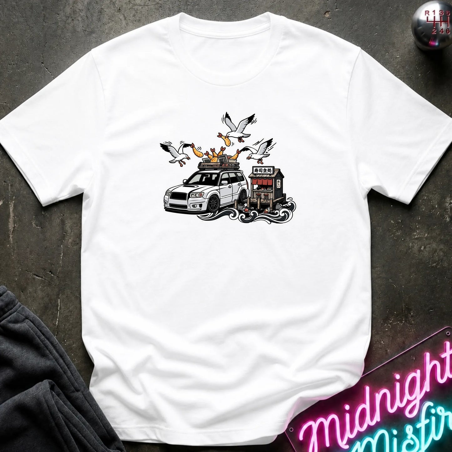 A white short-sleeve crewneck t-shirt with a graphic print of a white station wagon with a roof rack, seagulls, and a small building, laid flat on a dark textured surface.