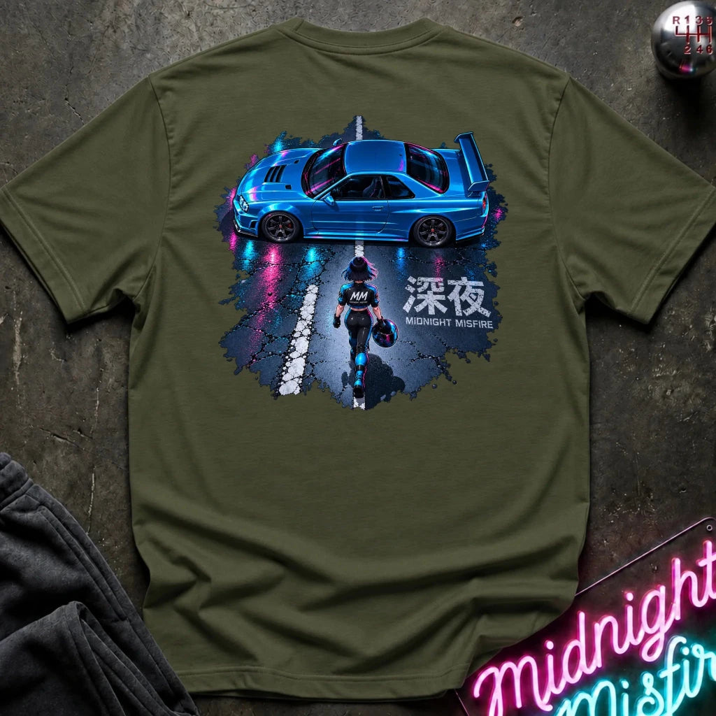 An olive green short-sleeve t-shirt laid flat on a dark textured surface, featuring a large graphic print on the back of a blue sports car and a driver with Japanese text. 