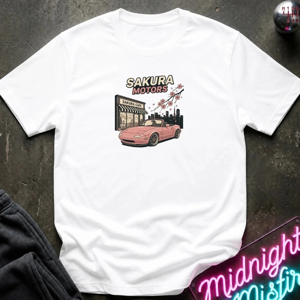 A white short-sleeve t-shirt laid flat on a dark textured surface, featuring a graphic print of a pink convertible car in front of a building with the text 'SAKURA MOTORS' and cherry blossoms. 