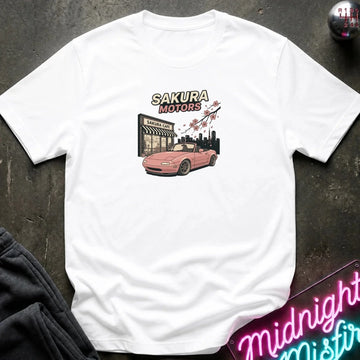 A white short-sleeve t-shirt laid flat on a dark textured surface, featuring a graphic print of a pink convertible car in front of a building with the text 'SAKURA MOTORS' and cherry blossoms. 