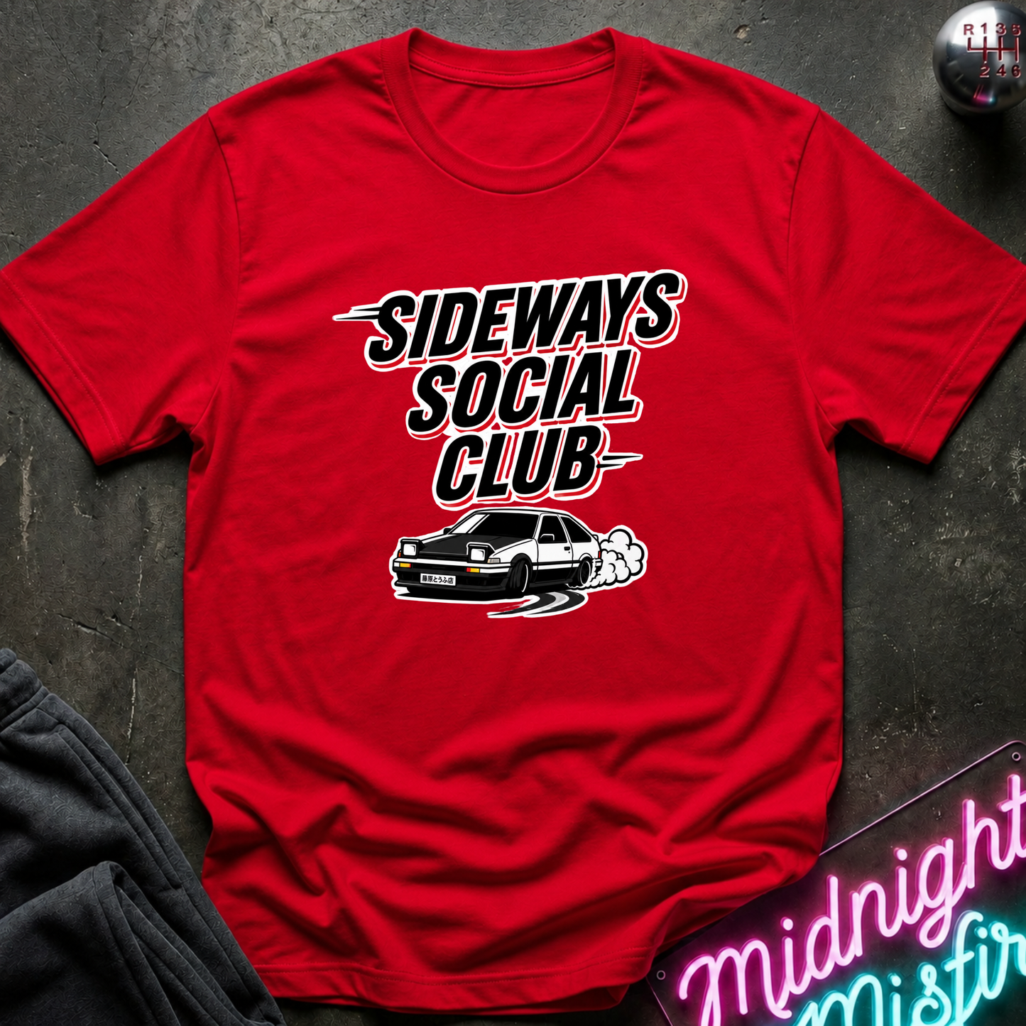 A red short-sleeve graphic T-shirt laid flat on a dark textured surface. The shirt features the text 'SIDEWAYS SOCIAL CLUB' in a stylized black-and-white font above a white car illustration with smoke.