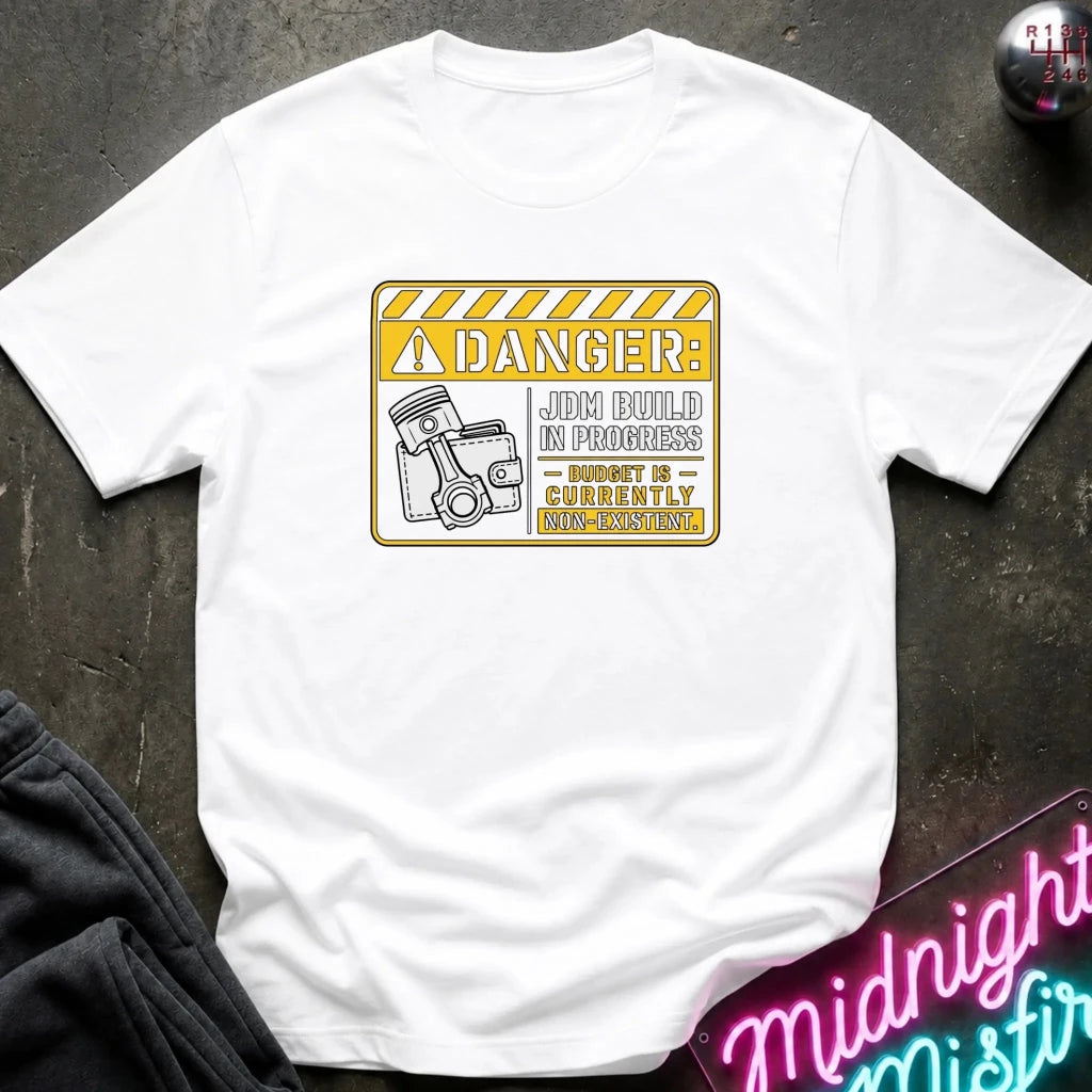 A white short-sleeve crewneck t-shirt laid flat on a dark textured surface. The shirt features a yellow and black warning sign graphic on the chest with the text 'DANGER: JOB BUILD IN PROGRESS - 