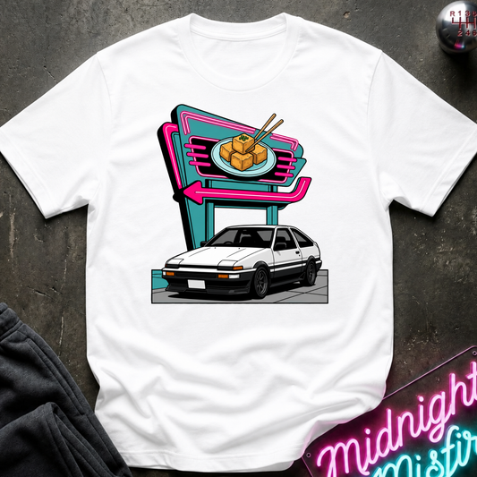 A white short-sleeve t-shirt laid flat on a dark textured surface, featuring a colorful graphic print of a white Toyota AE8 sports car parked in front of a neon-style sign with a plate of tofu and chopsticks.