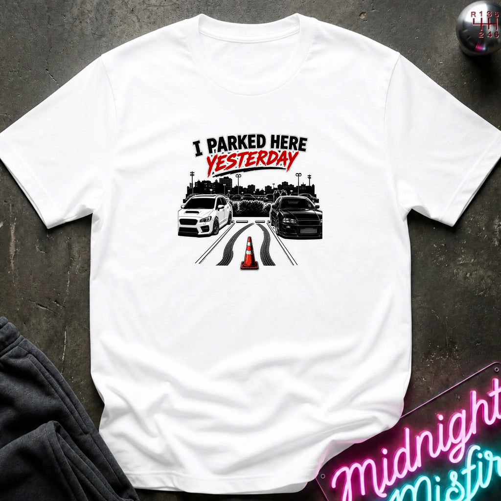 A white short-sleeved t-shirt  that has 2 cars parked in a space in between empty space with skid marks.