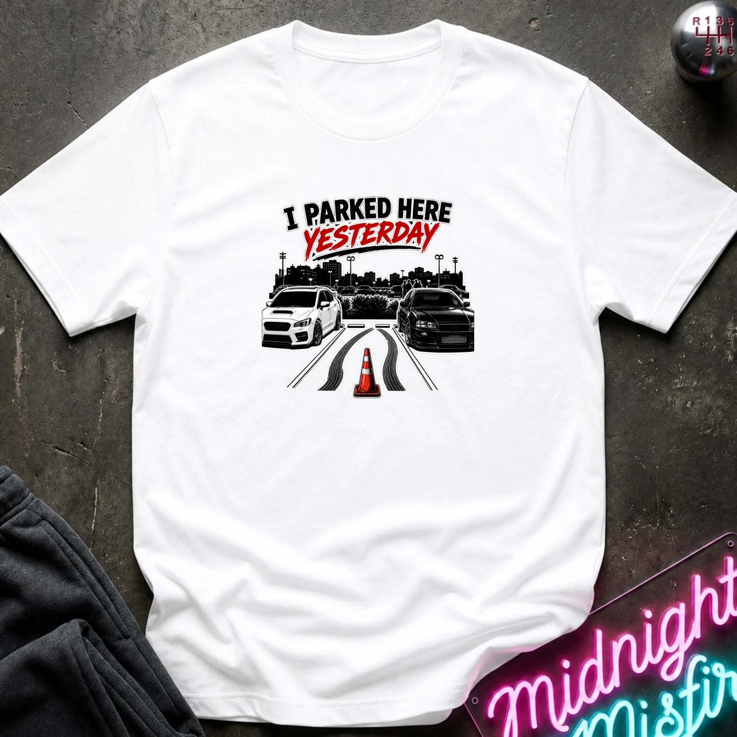 A white short-sleeved t-shirt  that has 2 cars parked in a space in between empty space with skid marks.