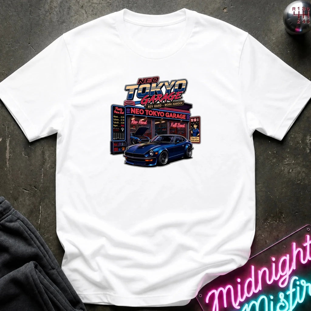 A white short-sleeve t-shirt laid flat on a dark textured surface, featuring a colorful graphic print of a blue sports car in front of a neon 'Neo Tokyo Garage' sign.