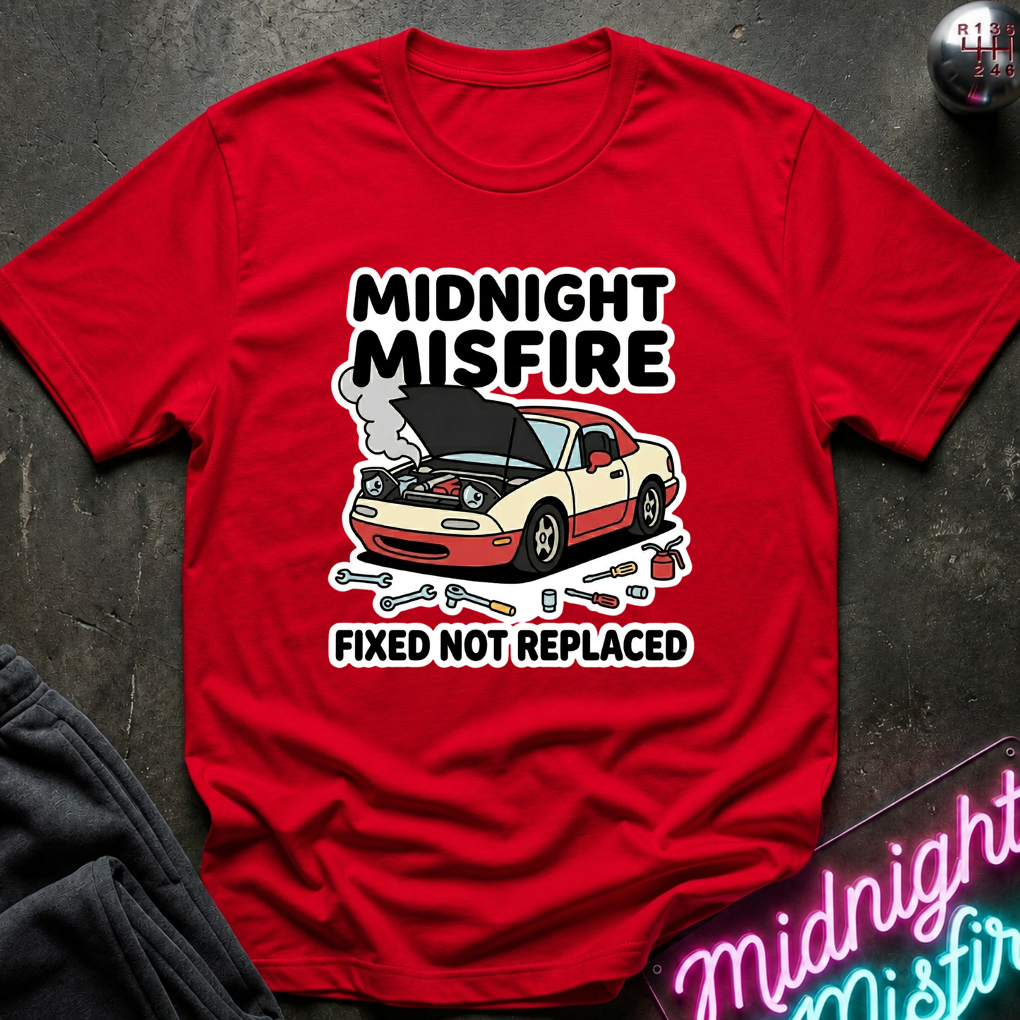 A red short-sleeve graphic T-shirt laid flat on a dark textured surface. The shirt features a cartoon car with its hood open, smoke, and the text 'MIDNIGHT MISFIRE' above and 'FIXED NOT REPLACED' below.
