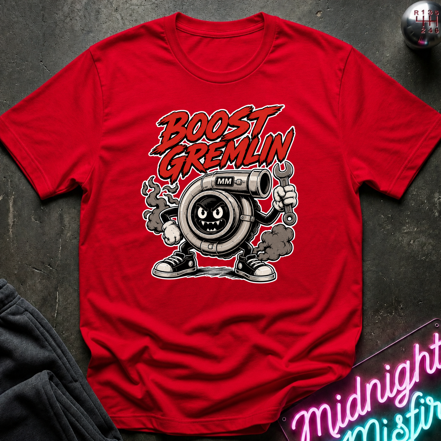 A red short-sleeve graphic T-shirt laid flat on a dark textured surface. The shirt features a cartoon turbocharger character with the text 'BOOST GREMLIN' above it.