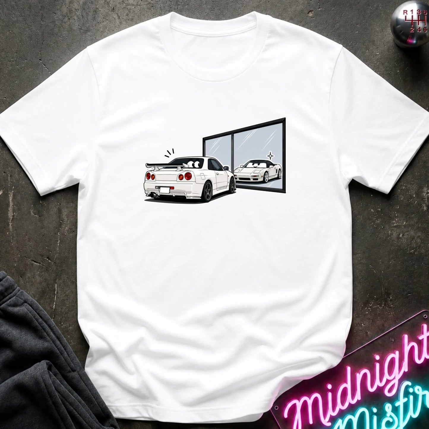 A white short-sleeve t-shirt laid flat on a dark textured surface, featuring a graphic print of a white sports car and its reflection in a window. A neon sign is partially visible in the bottom right corner.