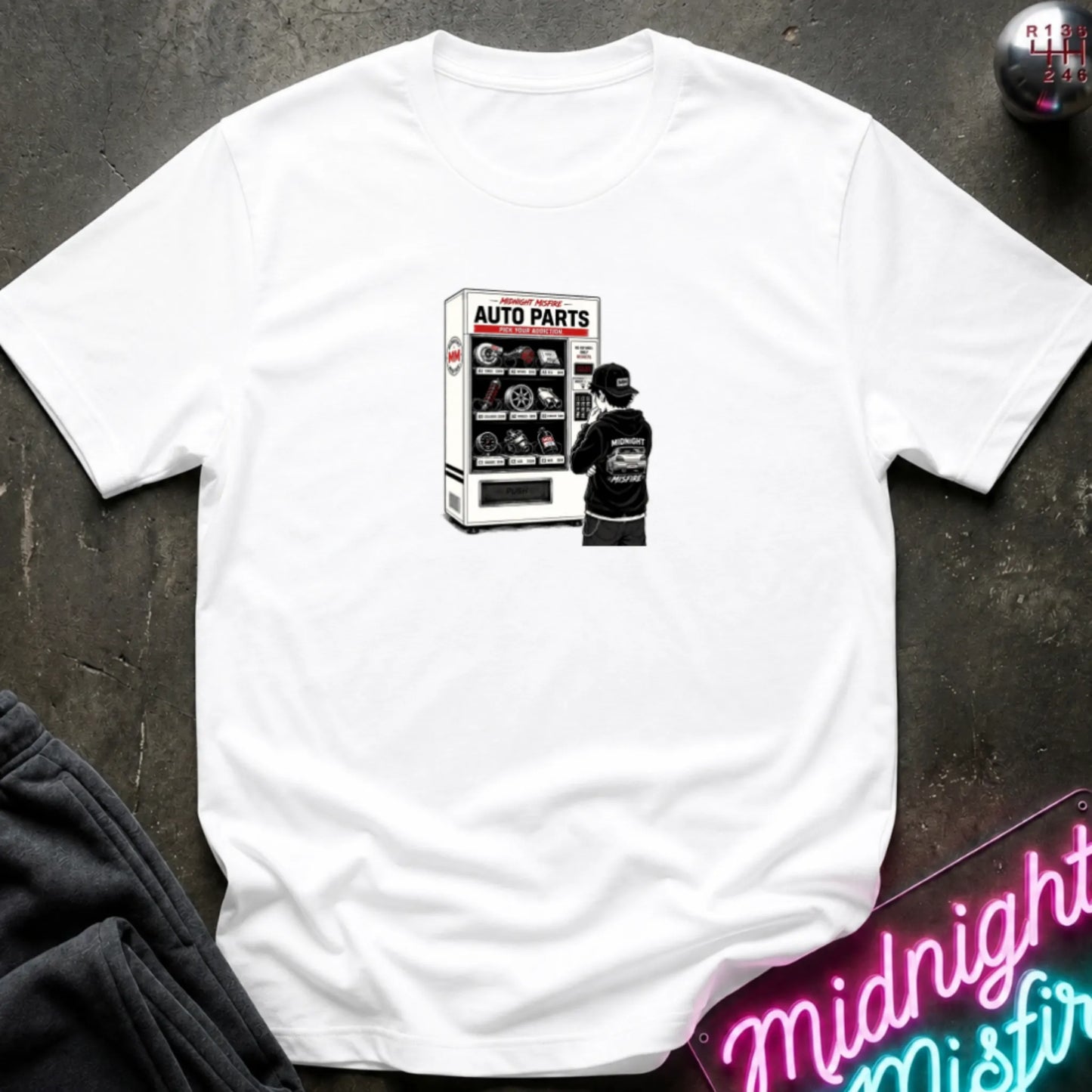 a white  short-sleeve t-shirt laid flat on a dark textured surface, featuring a graphic print of a vending machine and owner deciding.