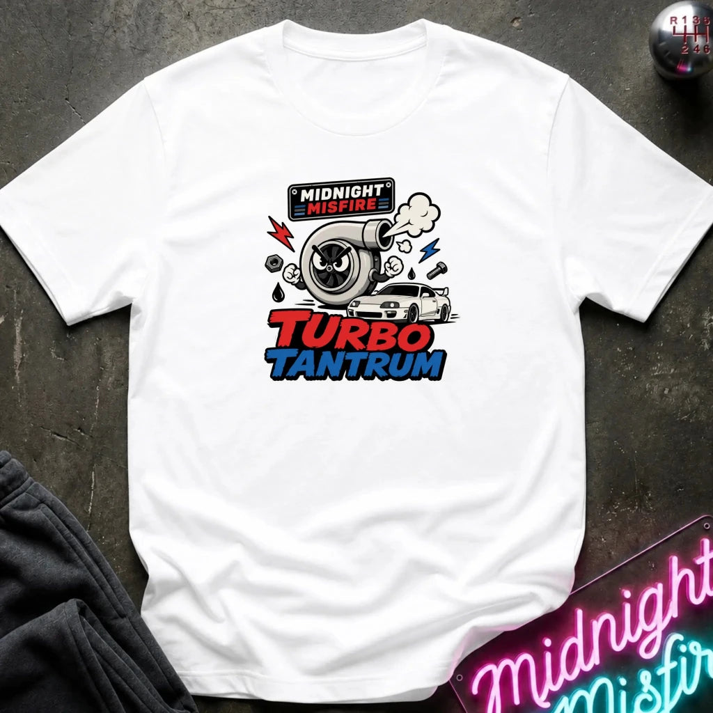 A white short-sleeve graphic t-shirt laid flat on a dark textured surface. The graphic features a turbocharger illustration with the text 'MIDNIGHT MISFIRE' above and 'TURBO TANTRUM' below in red and blue.