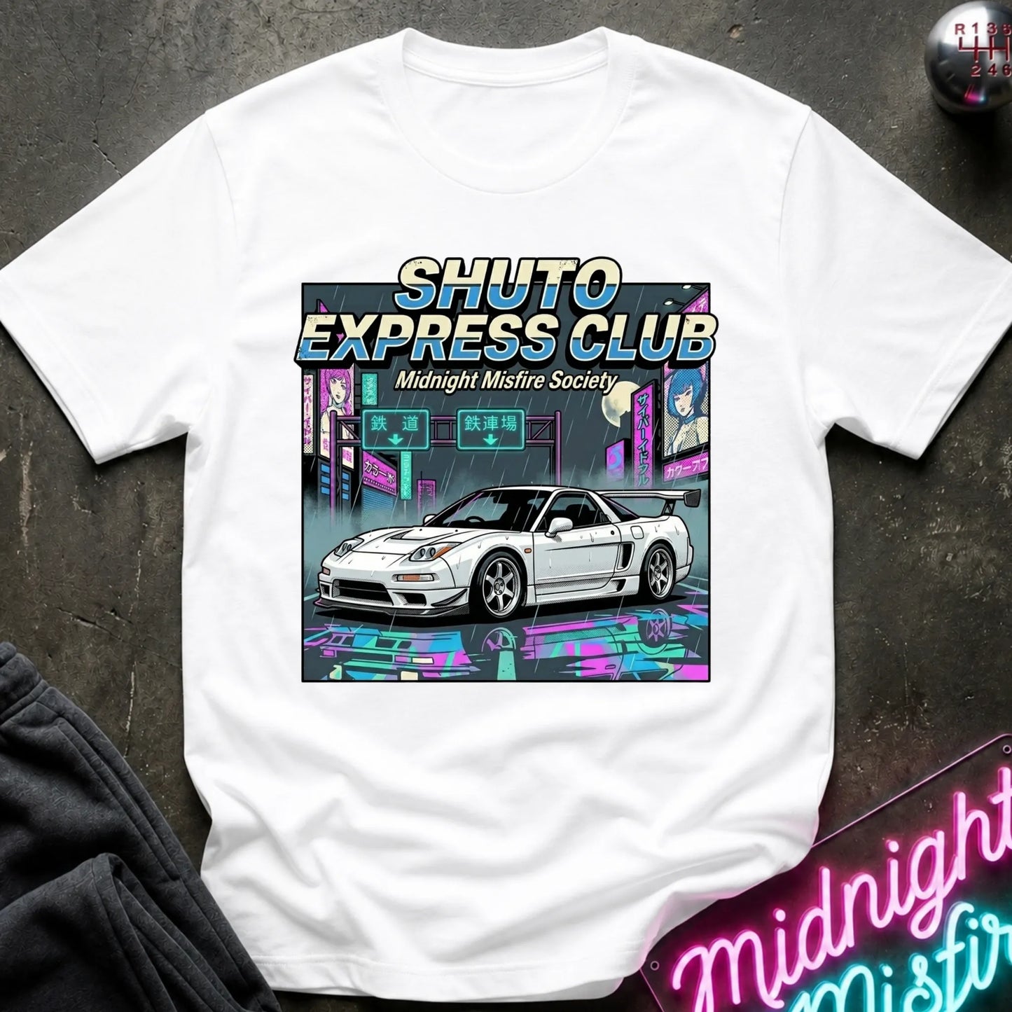 A white short-sleeve graphic T-shirt laid flat on a dark textured surface. The shirt features a large colorful front print with the text 'SHUTO EXPRESS CLUB' and 'Midnight Misfire Society' above an illustration of a white sports car in a neon cyberpunk city scene.