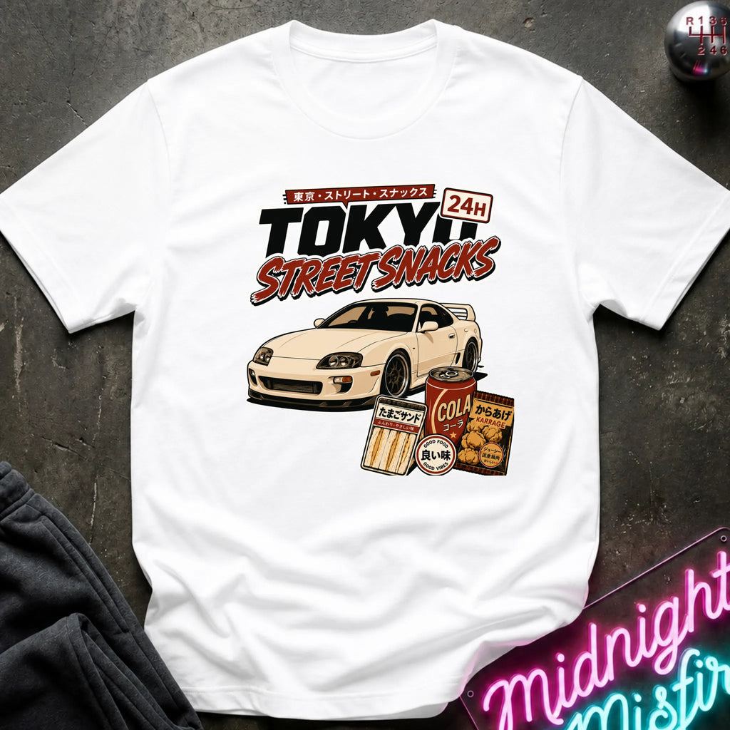A white graphic t-shirt laid flat on a dark textured surface. The graphic features a white sports car and retro Japanese-style snack cans with the text 'TOKYO STREET SNACKS 24H'.