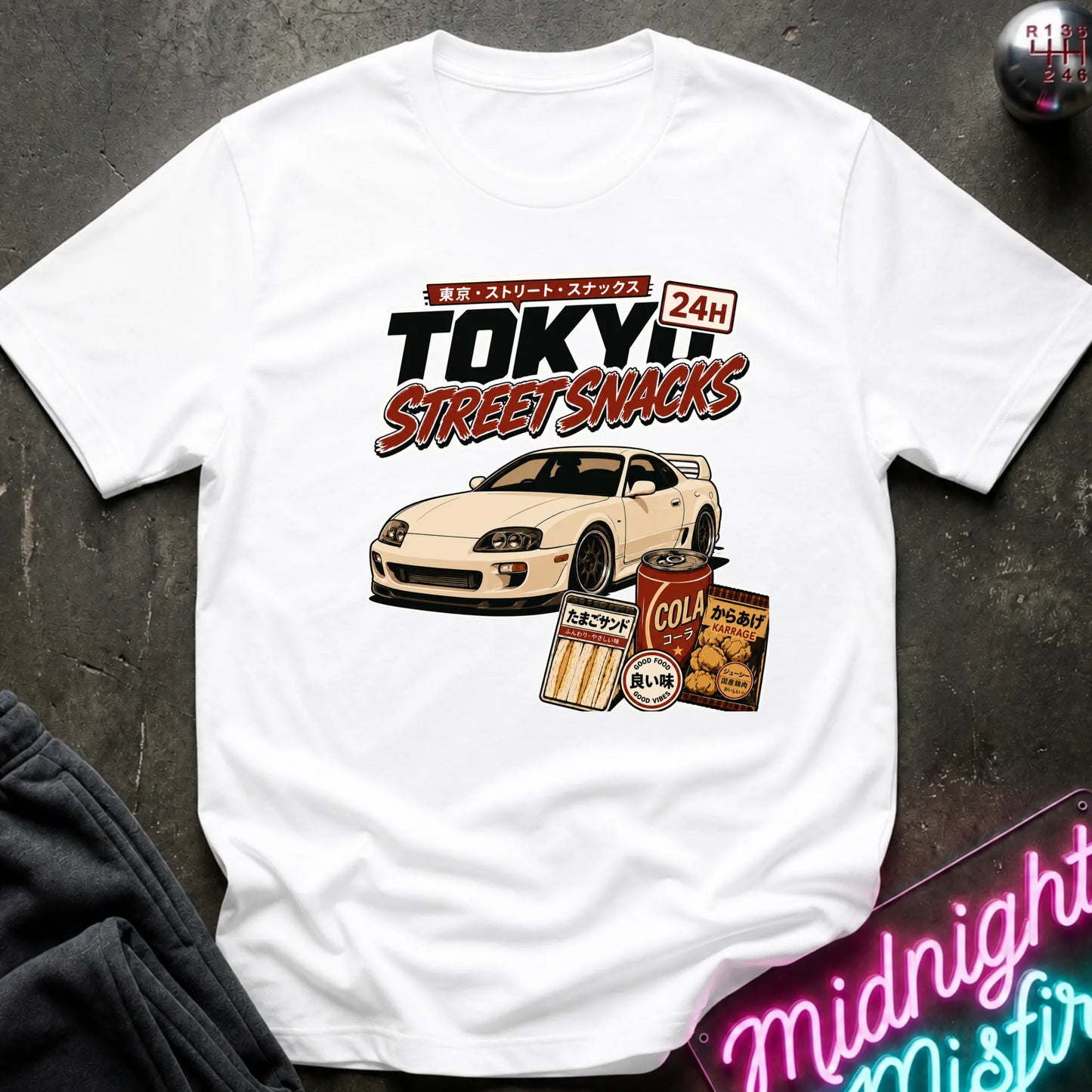 A white graphic t-shirt laid flat on a dark textured surface. The graphic features a white sports car and retro Japanese-style snack cans with the text 'TOKYO STREET SNACKS 24H'.