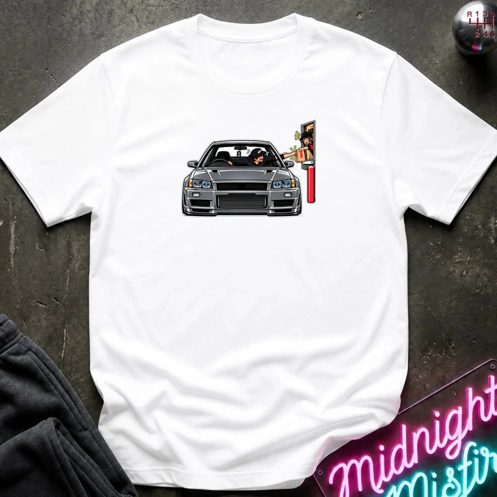 A white short-sleeve t-shirt laid flat on a dark textured surface, featuring a graphic print of a gray sports car with a driver and a red-and-yellow sign on the chest. 