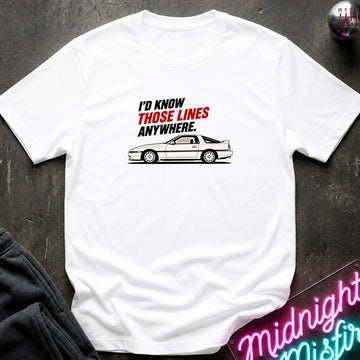 A white short-sleeve t-shirt laid flat on a dark textured surface, featuring a graphic of a white sports car and the text 'I'D KNOW THOSE LINES ANYWHERE.' in black and red.