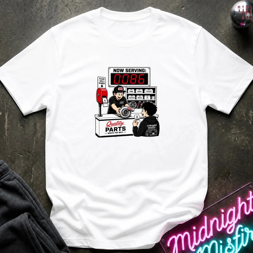 A white short-sleeve crewneck t-shirt featuring a retro-style graphic of a mechanic working at a parts counter with a digital display showing 'NOW SERVING: 0086'. 