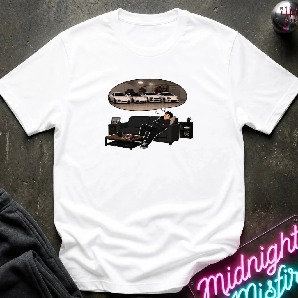 A white short-sleeve graphic t-shirt laid flat on a dark textured surface. The graphic shows a man sitting on a black couch dreaming, with an oval reflection of several white cars above him.