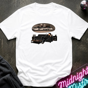 A white short-sleeve graphic t-shirt laid flat on a dark textured surface. The graphic shows a man sitting on a black couch dreaming, with an oval reflection of several white cars above him.