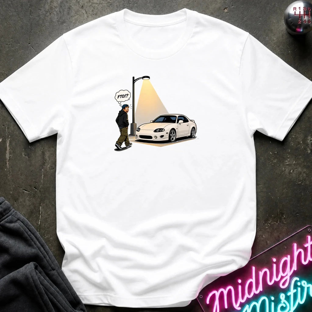 A white short-sleeve t-shirt laid flat on a dark textured surface, featuring a graphic print of a white sports car under a streetlamp with a speech bubble 