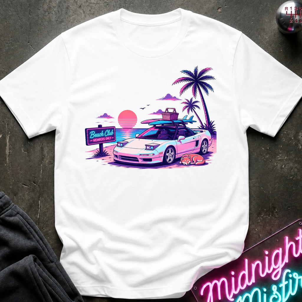 A white short-sleeve graphic t-shirt laid flat on a dark textured surface. The graphic features a retro-style illustration of a sports car with surfboards on its roof, next to palm trees, a sunset, and a 'Beach Club' sign.