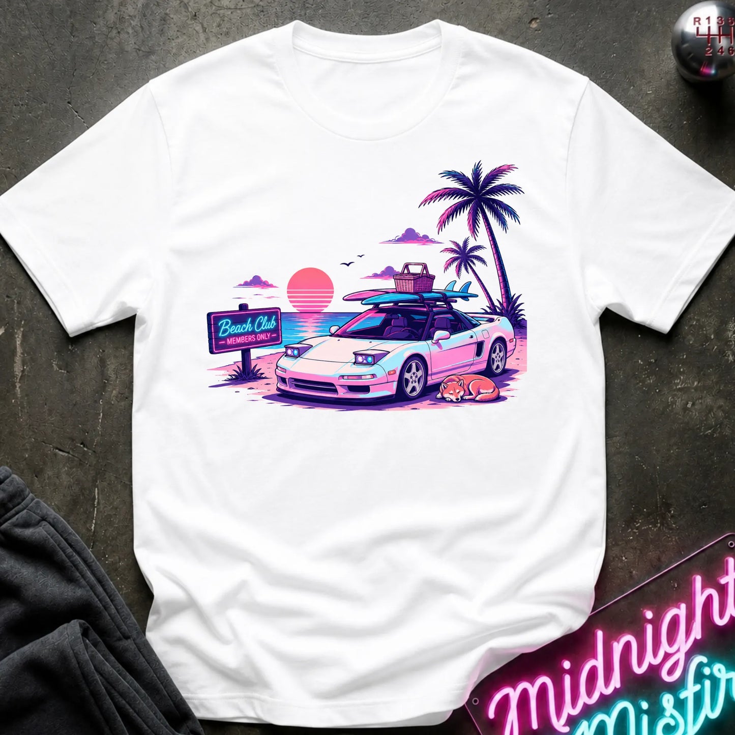A white short-sleeve graphic t-shirt laid flat on a dark textured surface. The graphic features a retro-style illustration of a sports car with surfboards on its roof, next to palm trees, a sunset, and a 'Beach Club' sign.
