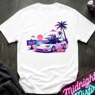 A white short-sleeve graphic t-shirt laid flat on a dark textured surface. The graphic features a retro-style illustration of a sports car with surfboards on its roof, next to palm trees, a sunset, and a 'Beach Club' sign.