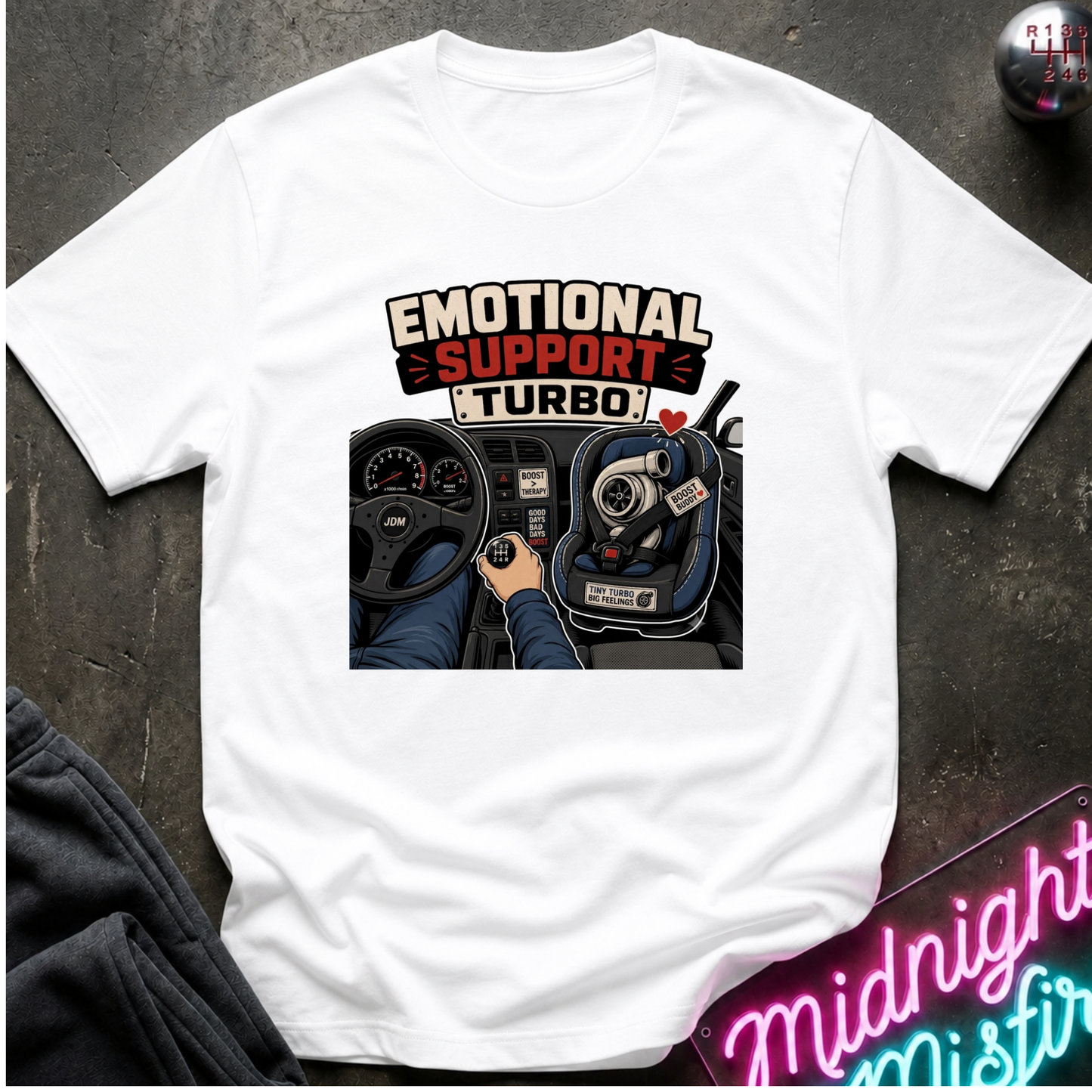 A white short-sleeve t-shirt laid flat on a dark textured surface, featuring a graphic print of a car dashboard and steering wheel with the text 'EMOTIONAL SUPPORT TURBO'.
