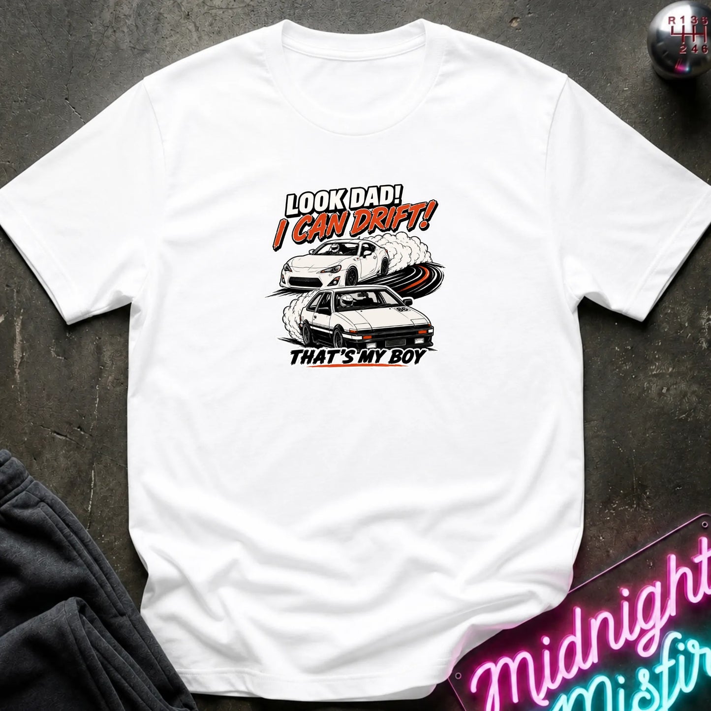A white short-sleeved t-shirt  that has a cars drifting which are older and newer generations of toyotas.