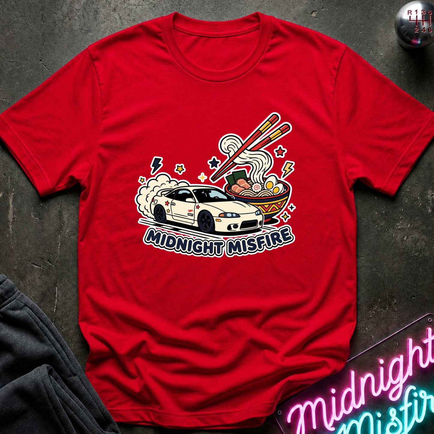 A red short-sleeve graphic T-shirt laid flat on a dark textured surface. The shirt features a colorful front print of a white sports car racing beside a bowl of ramen with chopsticks, plus the text 'MIDNIGHT MISFIRE.