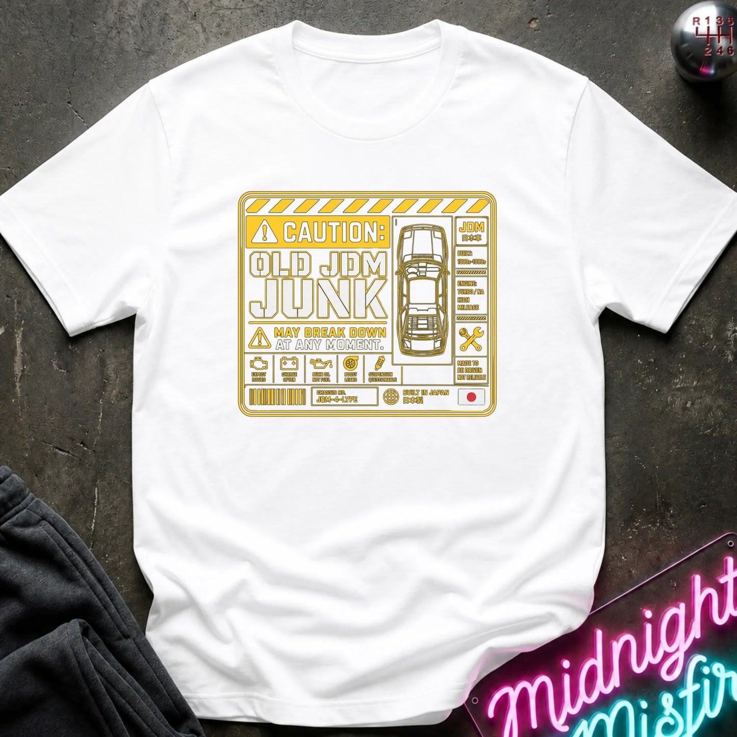 A white short-sleeve crewneck t-shirt with a yellow and black graphic print on the front. The graphic features a caution-style label with the text 'CAUTION: OLD JDM JUNK MAY BREAK DOWN' and a car illustration.