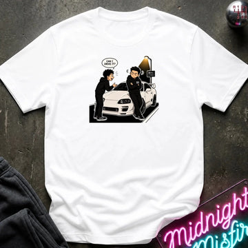 A white short-sleeve graphic t-shirt laid flat on a dark textured surface. The graphic shows a cartoon illustration of two people standing next to a white sports car, with one person holding the car door open and a speech bubble that reads 'CAN I DRIVE IT?'.