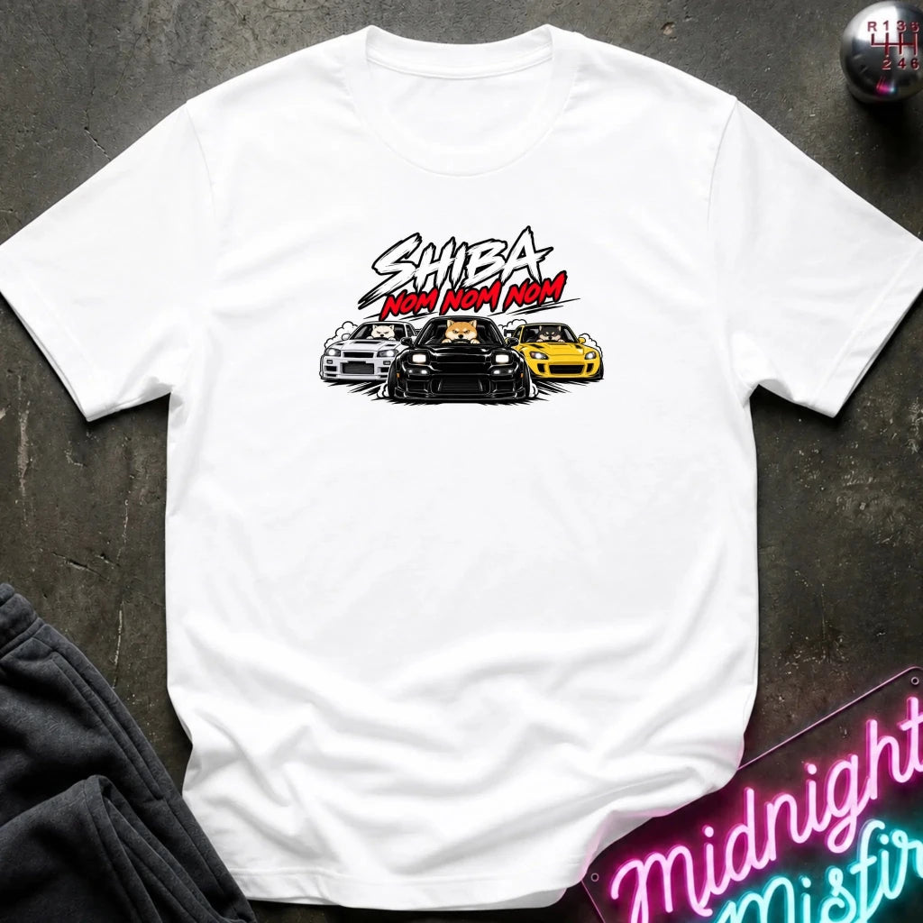 A white short-sleeve graphic t-shirt laid flat on a dark textured surface. The graphic features the text 'SHIBA NOM NOM NOM' in white and red, above an illustration of three cars (white, black, and yellow).