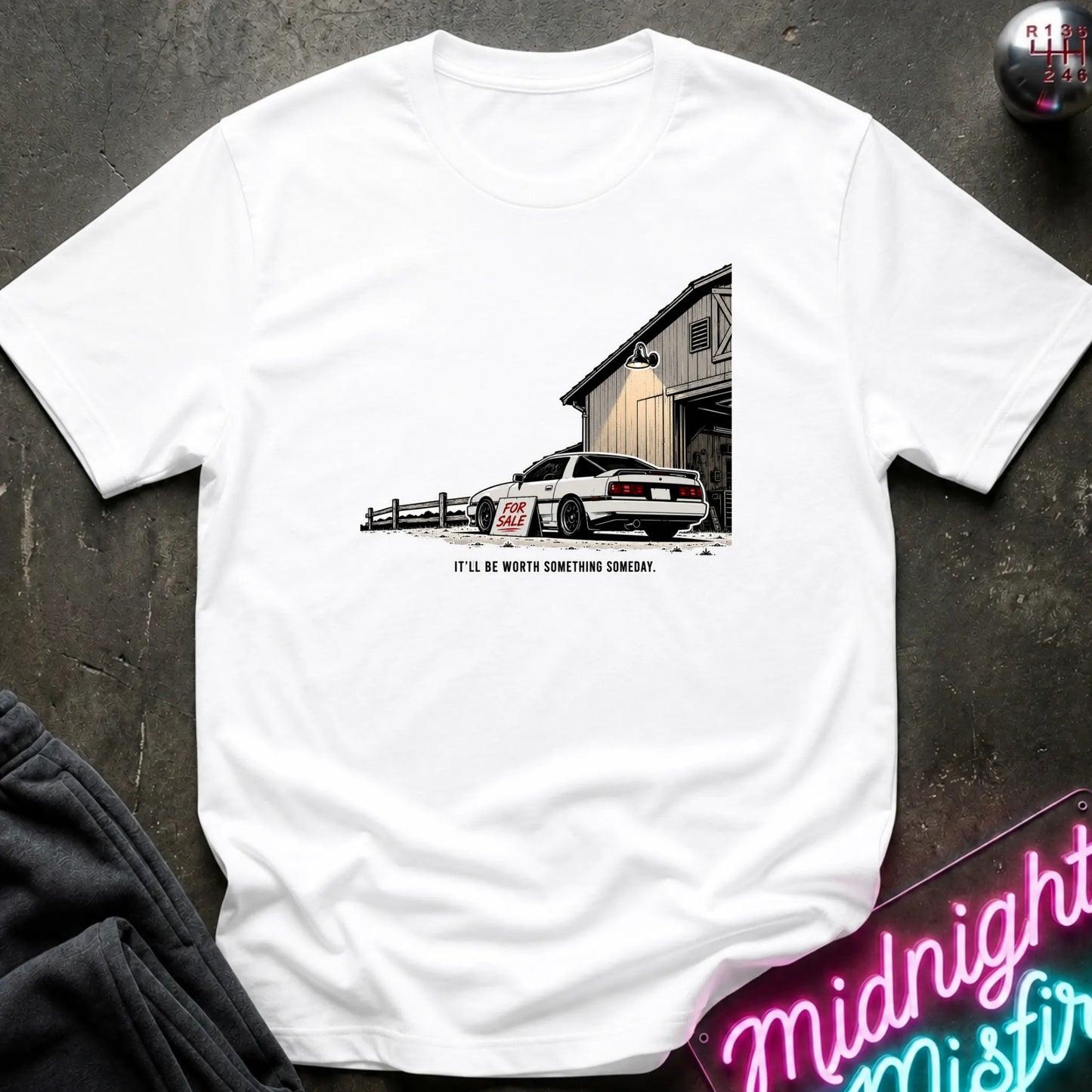 A white short-sleeve graphic t-shirt laid flat on a dark textured surface. The graphic shows a white sports car parked in front of a barn with the text 'IT'LL BE WORTH SOMETHING SOMEDAY.