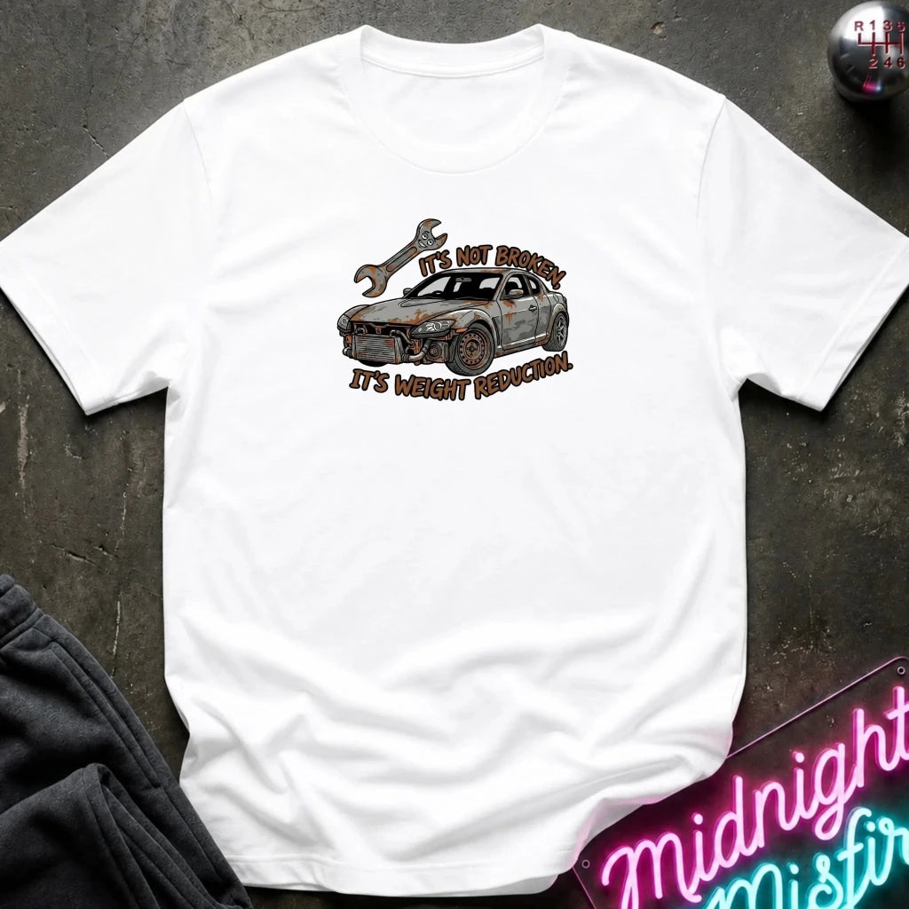 A white short-sleeve graphic t-shirt laid flat on a dark textured surface. The graphic shows a rusty car with a wrench and the text 'IT'S NOT BROKEN,